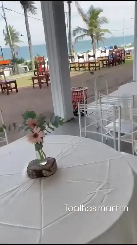 Quadro do vídeo com toalha marfim e arranjo de flores em uma mesa de festa à beira-mar